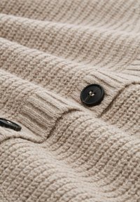 Tissu en tricot beige avec une texture côtelée ; présente des boutons noirs lisses ; le matériau doux tombe en douces vagues.