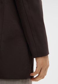 Donkerbruine blazer met een soepele textuur, met een getailleerde pasvorm en een licht gestructureerde schouder, gecombineerd met een hand in een neutrale positie.