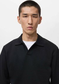 Joven con el cabello negro corto lleva una camisa negra de cuello sobre una camiseta blanca frente a un fondo blanco liso.