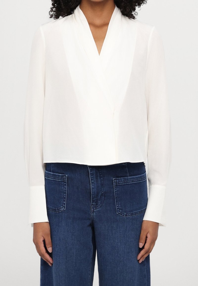 Femme portant un chemisier blanc à manches longues avec de larges poignets et un jean bleu taille haute avec des poches plaquées à l'avant.