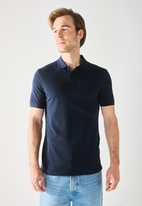 Polo bleu marine avec col, manches courtes et patte de boutonnage à deux boutons. Tissu lisse ; associé à un denim bleu clair.