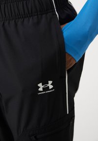 Melnas sporta bikses ar elastīgu jostasvietu, baltām akcentiem un ar rāvējslēdzēju aizveramu kabatu. Uz kājas ir sudraba Under Armour emblēma.