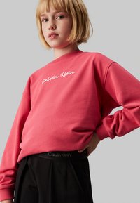 Felpa rosa con colletto rotondo e maniche lunghe, con logo "Calvin Klein" ricamato in bianco sul petto, abbinata a pantaloni neri.