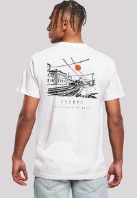 Mann mit geflochtenem Haar, der ein weißes T-Shirt mit schwarzem Sketch von Eisenbahnschienen, Gebäuden und einer orangefarbenen Sonne auf dem Rücken trägt.