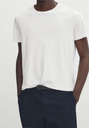 Homme portant un t-shirt blanc uni à manches courtes, rentré dans un pantalon bleu marine foncé avec une ceinture noire, debout les mains dans les poches.