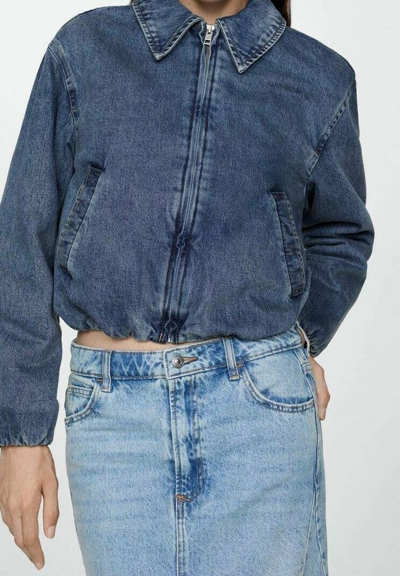 Personne portant une veste courte en denim bleu foncé zippée avec un ourlet élastique et une jupe en denim bleu clair taille haute avec bouton et fermeture éclair.