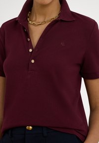 Κεραμιδί κοντομάνικο πόλο μπλουζάκι με γιακά, τρία χρυσά κουμπιά και μικρό κεντητό λογότυπο στο στήθος. Λείο ύφασμα.