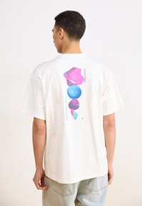 Kortærmet creme farvet t-shirt med et farverigt abstrakt design på ryggen i pink og blå, med grafisk tekst og en afslappet pasform.