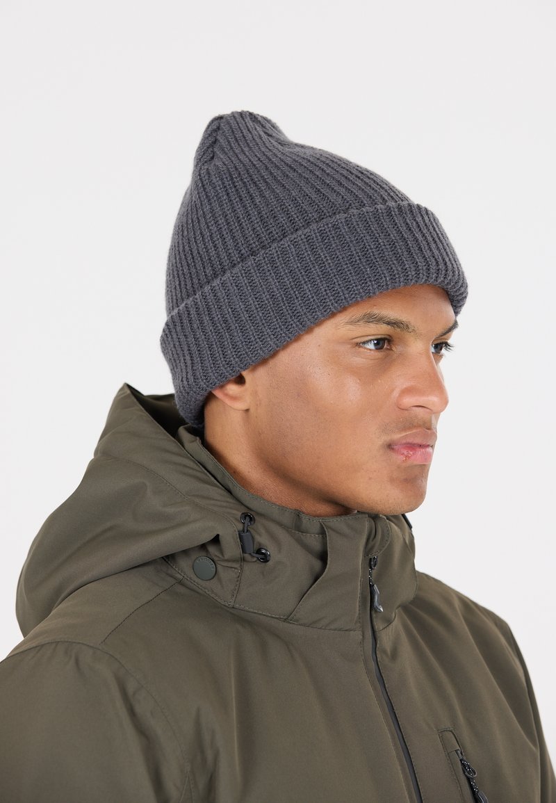 Grijze geribde gebreide beanie met een puntige top, gedragen met een olijfgroene hooded jacket met trekkoorden en ritssluitingzakken.