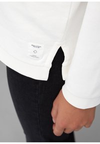 Camicia bianca a maniche lunghe con uno spacco laterale e un'etichetta di tessuto sull'orlo, abbinata a pantaloni in denim scuri. Texture liscia, design casual.