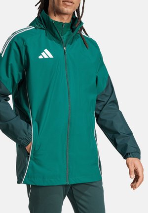 Groene windjack met witte accenten, ritsluiting, hoge kraag en elastische boorden. Bevat een logo op de borst en zijzakken.