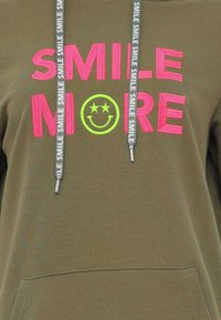 Felpa verde oliva con testo ricamato in rosa e giallo neon "SORRIDI DI PIÙ" e faccina sorridente. Include coulisse grigia con testo ripetuto.