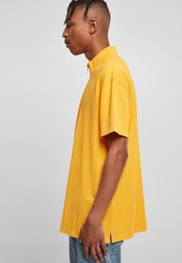 Gul kortermet polo shirt med ståkrage, mykt bomullsmateriale, avslappet passform, sidespalter og en glatt tekstur. Brukt med lyseblå jeans.