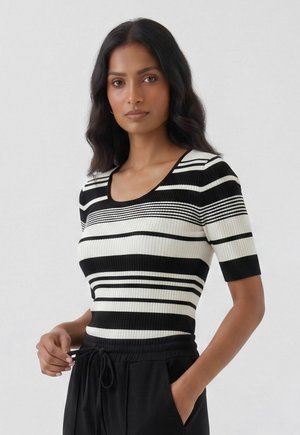 Woman with wavy black hair wearing a black and white striped short-sleeve top and black pants, standing against a plain light background.