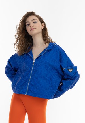 Modrá bomber bunda se vzorem, s předním zipem, kapucí a bočním zipovým kapsou; spárovaná s oranžovými legíny. Výrazná lesklá textura a akcenty.