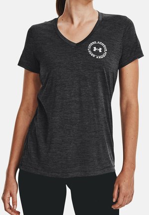 Vrouw die een donkergrijs V-hals T-shirt met korte mouwen van Under Armour draagt met een logo op de linkerborst, gecombineerd met een zwarte broek.