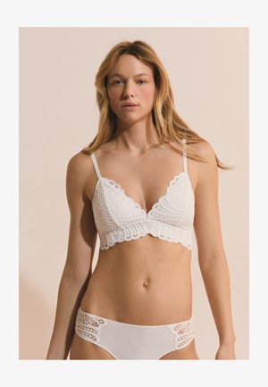 Donna che indossa un bralette bianco in pizzo e biancheria intima abbinata con dettagli in pizzo, in piedi davanti a uno sfondo beige, rivolta in avanti.