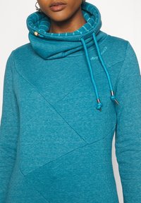 Sudadera turquesa con cuello con capucha, forro a rayas en contraste, cierre con cordón y detalles de panel diagonal texturizado.