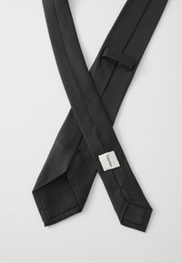 Melnā kaklasaite ar teksturētu apdari, kurai ir diagonāls raksts un kvadrātveida etiķete ar uzrakstu "Calvin Klein" vienā pusē.