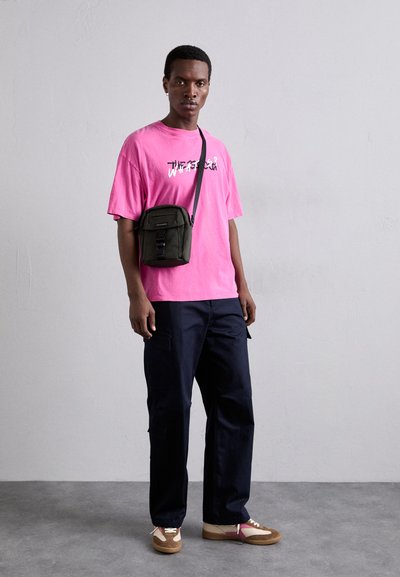 Růžové oversized tričko s bílou grafikou, navy cargo kalhoty a hnědé tenisky s růžovými akcenty. Zelená crossbody taška se šňůrkami.
