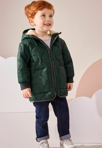 Grön quiltad jacka med huva, texturerat mönster, dragkedjestängning och två fickor fram, bärs över en grå skjorta och mörka jeans.