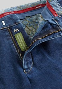 Donkerblauwe jeans met gedeeltelijk open ritssluiting, gedessineerde binnenvoering en een groene label met de tekst "pure organische katoenstof, sociaal verantwoord gemaakt."