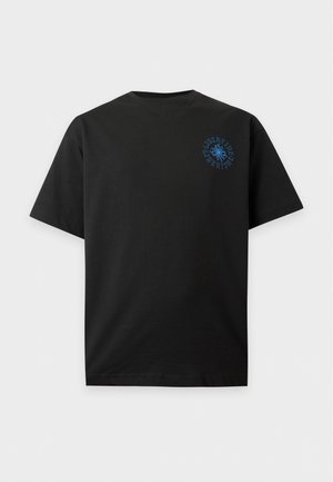 Homme portant un t-shirt noir avec un logo bleu sur la poitrine, un jean large bleu clair et des chaussures marron, debout avec une main dans la poche.