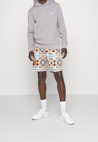 Sudadera gris con logo, pantalones cortos estampados con diamantes en naranja, azul y blanco, zapatillas blancas y calcetines de longitud media.