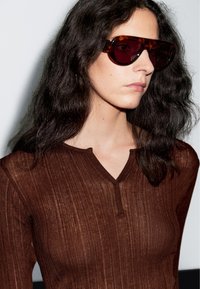 Braunes, geripptes Langarmoberteil mit V-Ausschnitt und Knopfleiste. Das Model trägt eine dunkle, übergroße Sonnenbrille, das Haar ist wellig und dunkel. Der Hintergrund ist hell.