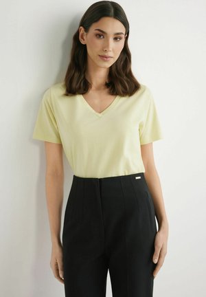 Kobieta o ciemnych włosach do ramion, ubrana w jasnżółtą bluzkę z dekoltem w serek i czarne spodnie z wysokim stanem, stoi przy białej ścianie.