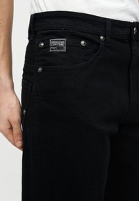 Κοντινό πλάνο ενός ατόμου που φοράει μαύρο τζιν παντελόνι Versace Jeans Couture με μεταλλικά καρφιά και ορατή ετικέτα μάρκας στην μπροστινή τσέπη.