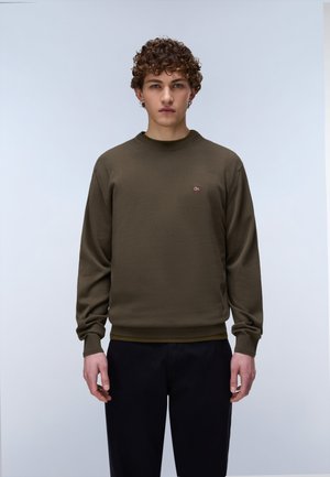 Jeune personne aux cheveux bouclés portant un sweat-shirt vert olive et un pantalon noir, debout devant un fond clair uni.