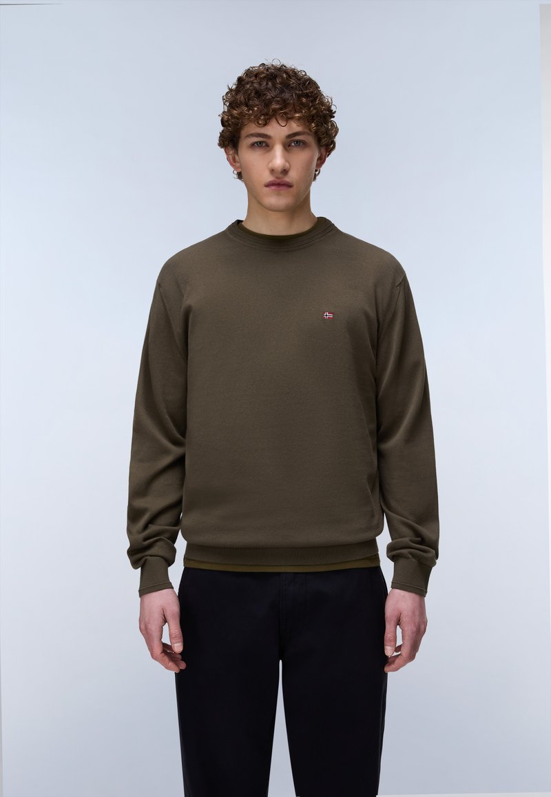 Jeune personne aux cheveux bouclés portant un sweat-shirt vert olive et un pantalon noir, debout devant un fond clair uni.