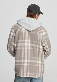 Koszula w kratę w odcieniach beżu i kremu, z szarym kapturem pod spodem. Posiada kołnierz i luźny krój.