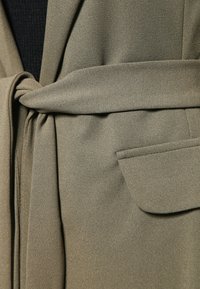 Abrigo envolvente verde oliva con una tela suave y texturizada, un cinturón atado en la cintura y un bolsillo en la parte delantera.