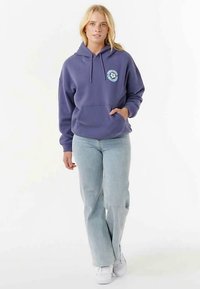 Lila Kapuzenpullover mit Fronttasche und Kordelzug an der Kapuze, mit einem runden Logo auf der linken Brust, kombiniert mit hellblauen Jeans.
