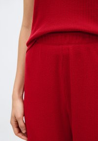 Κοντινό πλάνο ενός ατόμου που φοράει κόκκινο πλεκτό παντελόνι ριμπ και το αντίστοιχο τοπ, εμφανίζοντας τη μέση και μέρος του αριστερού βραχίονα και χεριού.