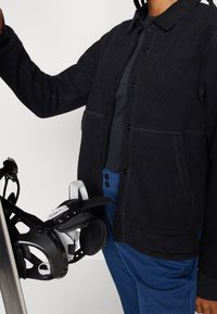 Giacca in pile nera con chiusura a bottoni, indossata sopra un top scuro. Pantaloni blu con due bottoni. Bindings da snowboard neri tenuti in una mano.