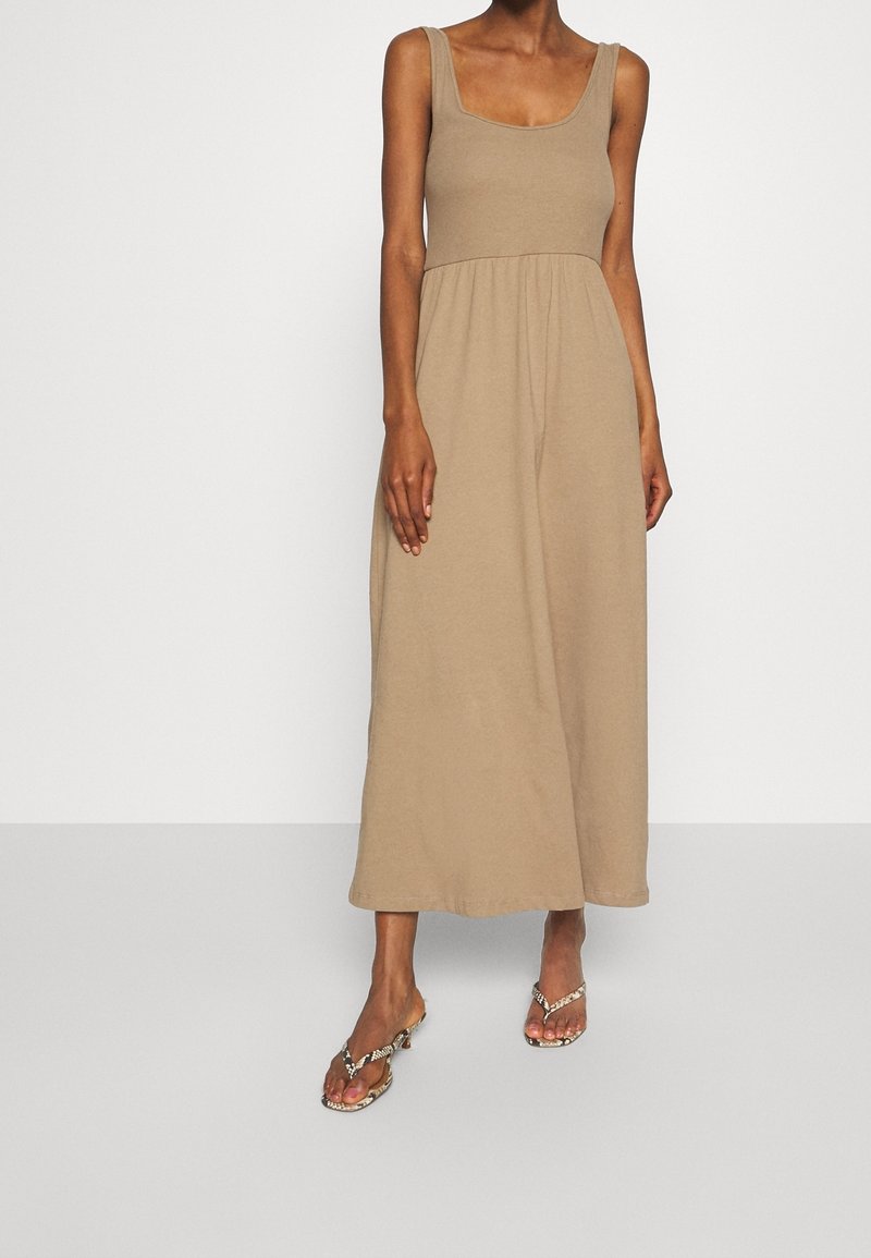 Femme portant une robe longue beige sans manches et des sandales à talons ouvertes avec un motif peau de serpent, debout devant un fond uni.
