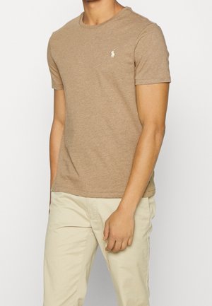 Mann trägt beige Kurzarm-T-Shirt mit kleinem weißen Logo auf der Brust und hellfarbige Hose, steht mit einem entspannten Arm.