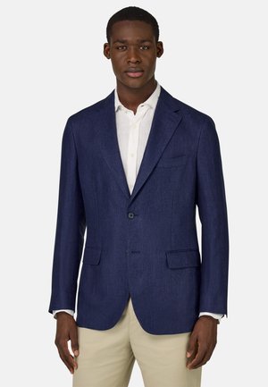 Blazer blu navy realizzato in tessuto leggero, caratterizzato da rever a punta, due tasche frontali e chiusura a un bottone. Indossato su una camicia bianca.