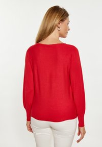 Roter Strickpullover mit langen Ärmeln, gerippten Bündchen und Saum, hat einen weiten Ausschnitt und eine lockere Passform. Getragen mit weißen Hosen.
