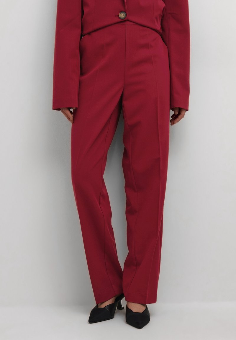 Femme portant un pantalon taille haute rouge sur mesure et un blazer assorti à bouton unique, avec des talons noirs à bouts pointus.