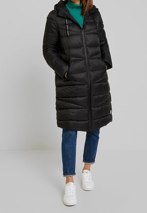 Zwarte pufferjas met capuchon, gequilt ontwerp, ritssluiting en twee voorkruiszakken, gedragen met blauwe jeans en witte sneakers.