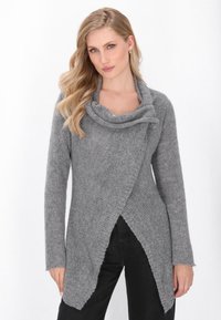 Donna con lunghi capelli biondi che indossa un cardigan grigio lavorato a maglia con un colletto drappeggiato e pantaloni neri, in piedi contro uno sfondo bianco.