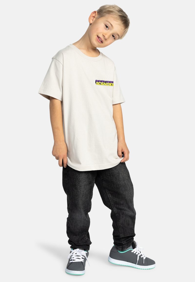 Světlé béžové oversized tričko s vícebarevným grafikou na levé straně hrudi. Nosené s tmavými džíny a šedými teniskami s bílými tkaničkami.