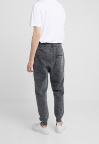 Man klädd i urblekta svarta joggingbyxor, vita sneakers och en enkel vit t-shirt, står framför en enfärgad ljusgrå bakgrund.