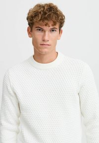 Pull en maille blanche avec un motif texturé, encolure ronde côtelée et manches longues ; finition lisse et matériau léger.