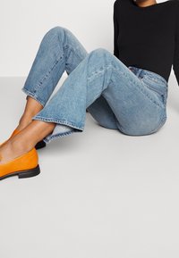 Jeans de mezclilla azul claro acampanados con dobladillo sin terminar, combinados con unos zapatos de ante naranja tipo slip-on con suela negra. Se puede ver una camiseta negra de manga larga.