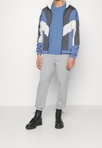 Osoba ubrana w niebiesko-czarną kurtkę w kolorowe bloki, niebieską koszulę, jasnoszare spodnie dresowe i czarne buty na sznurowadła stoi na tle białego tła.